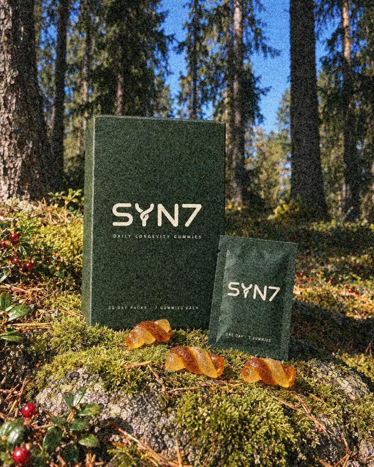 SYN7 box, a daily sachet and three twist gummies on a mossy boulder in a sunny pine forest