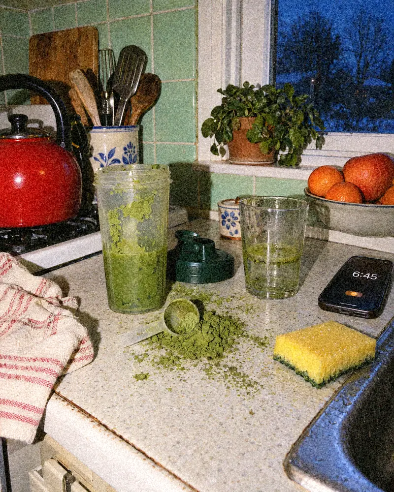 A kitchen counter at 06:45 with a green powder spill, a dirty shaker, a scoop and a sponge next to the sink